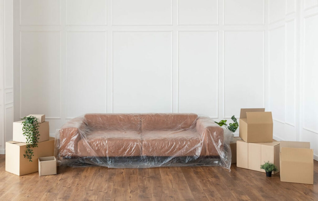 Brown couch with plastic cover sits in an empty living room surrounded by packed boxes.